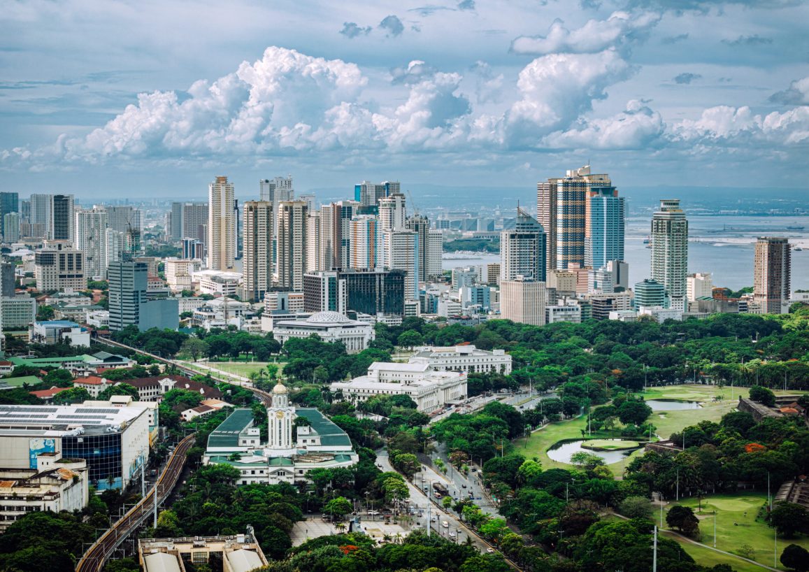 The image showcases Manila's vibrant urban landscape, including skyscrapers, green parks, and historical structures, all set under a beautiful cloudy sky.